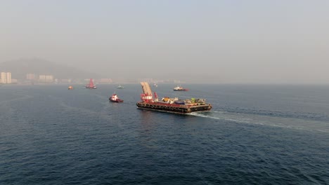 Barge-loaded-with-Concrete-mixer-trucks-pulled-to-port-by-a-Tugboat-in-Hong-Kong-bay,-Aerial-view