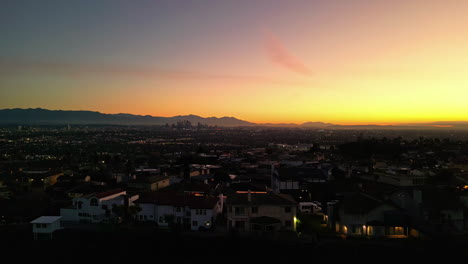 Aerial-view-backwards-over-a-neighborhood-with-Los-Angeles-dusk-skyline-background