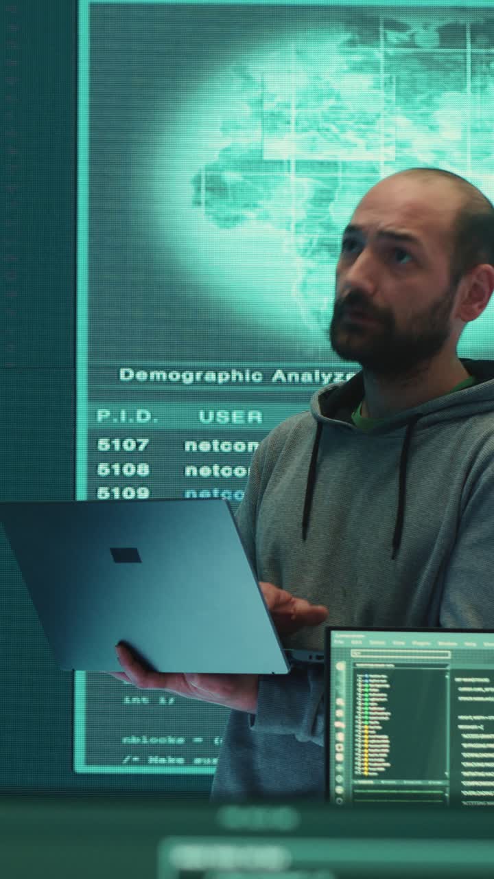 Male Hacker Working on Laptop in Dark Room with Multiple Screens