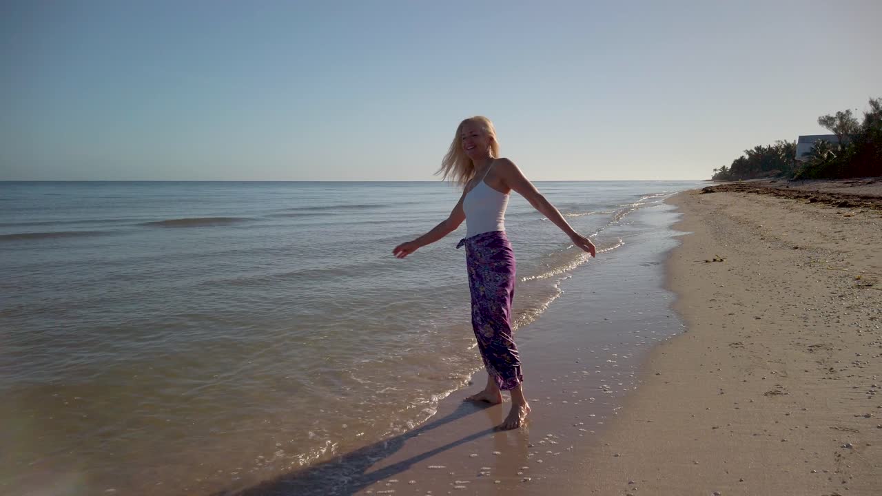 Slow motion of joyous mature woman backlit, splashing in the water on a beach at sunrise or sunset