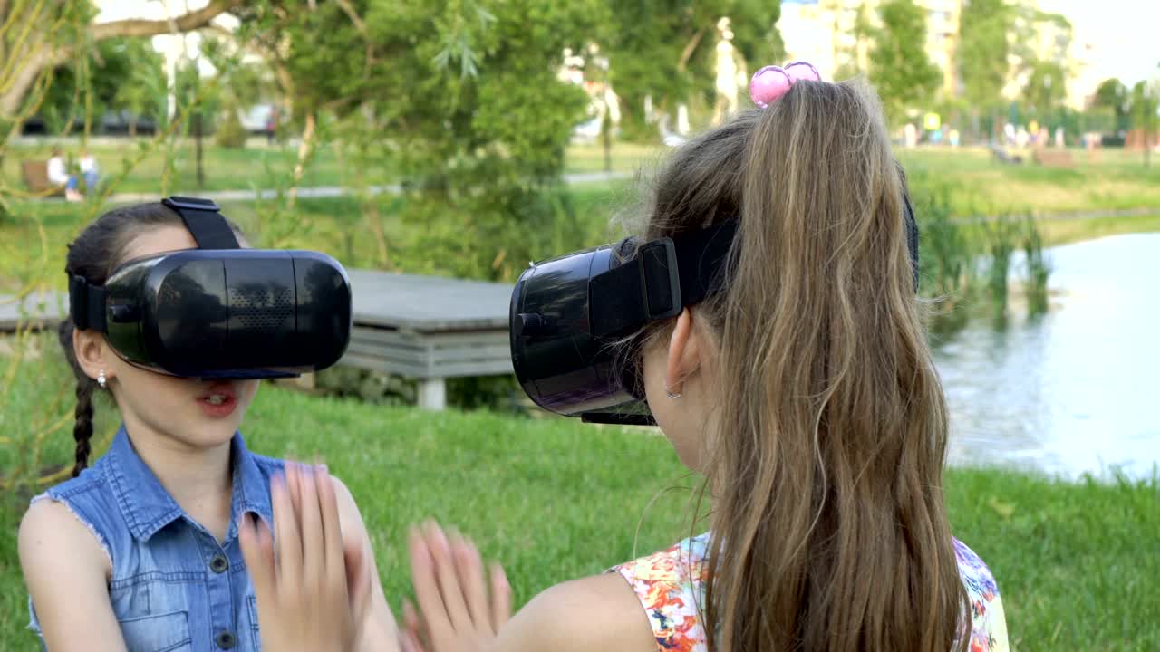 가상현실 안경을 입은 두 어린 소녀가 은 날 강변의 잔디에 앉아서 놀고 있습니다.