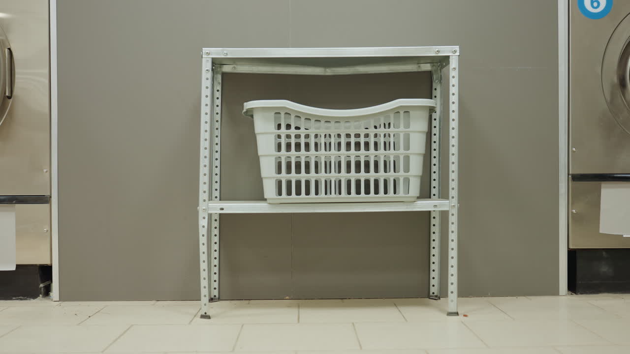 Close zoom on laundry basket on metal shelf between two industrial front load washers in clean laundromat, one door open, tiled floor and grey wall background, interior showing equipment symmetry