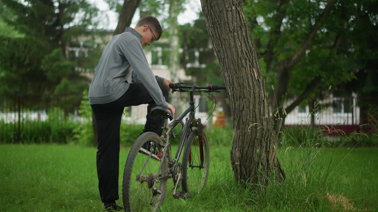 잔디로 된 공원에서 나무 에 주차 된 자전거를 고있는 소년의 뒷면, 소년은 좌석과 버에 손을 놓고 다리를 낮추기 전에 페달에 발을 집중합니다.
