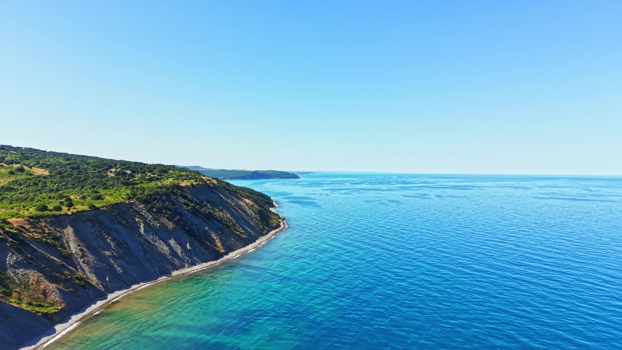 Onde che toccano la riva di un mare azzurro e limpido accanto a una collina verdeggiante in una giornata di sole in una zona costiera.