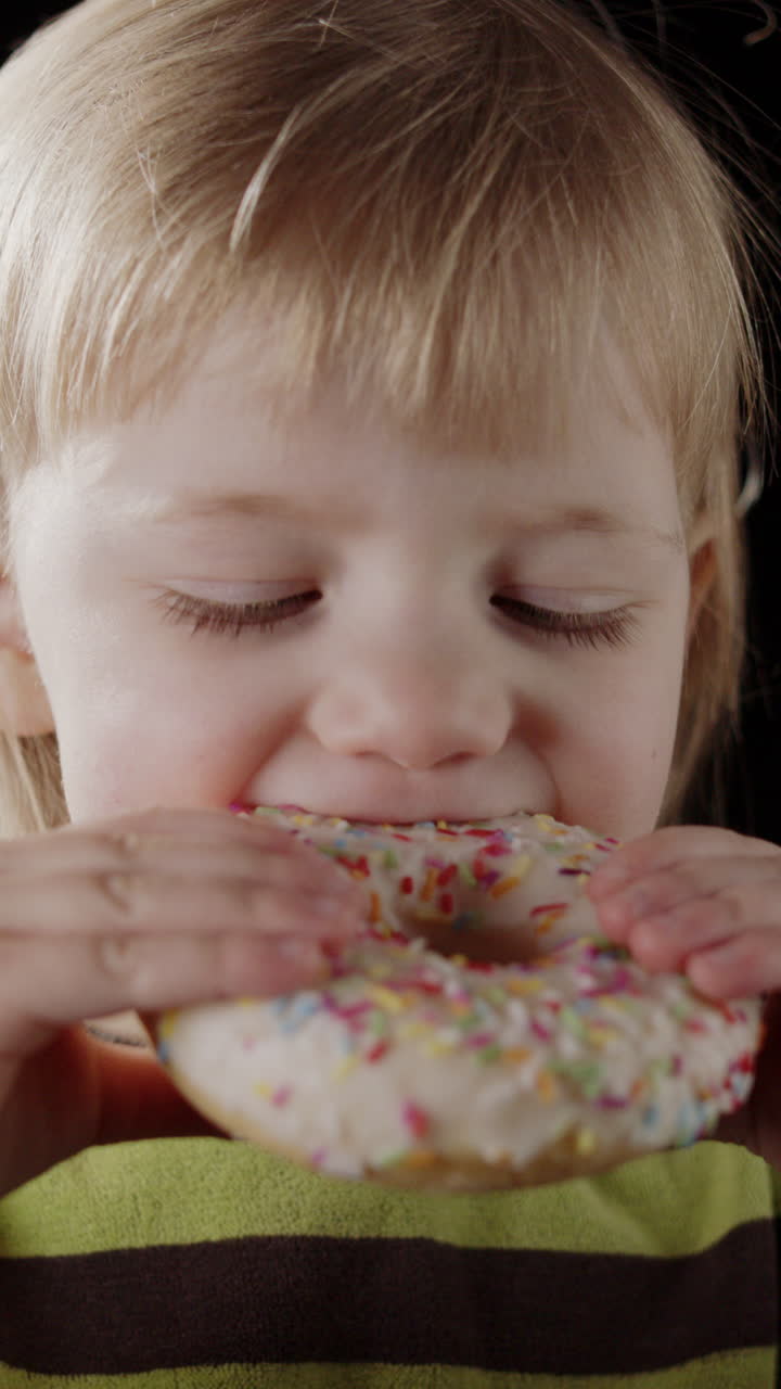 una chica adorable toma un gran bocado de rosquilla esmaltada con salpicaduras, frontal vertical
