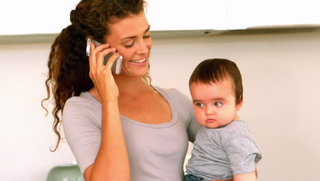 mother holding her baby son in the kitchen talking on phone