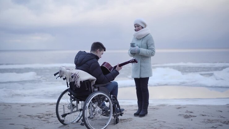 Young Caucasian disabled man playing guitar on river bank for mother or invalid tender. Boy in wheelchair enjoying autumn day outdoors. Lifestyle, hobby, disability, skill.