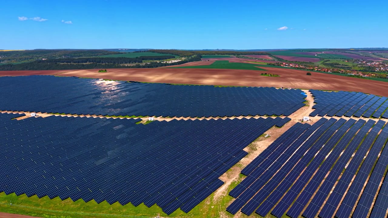 Store rækker af solcelleanlæg med fjern landsby og landbrugsjord. Forhøjet vinkel over skråtstillede paneler, der møder åben jord, spredte huse og skov i horisonten.