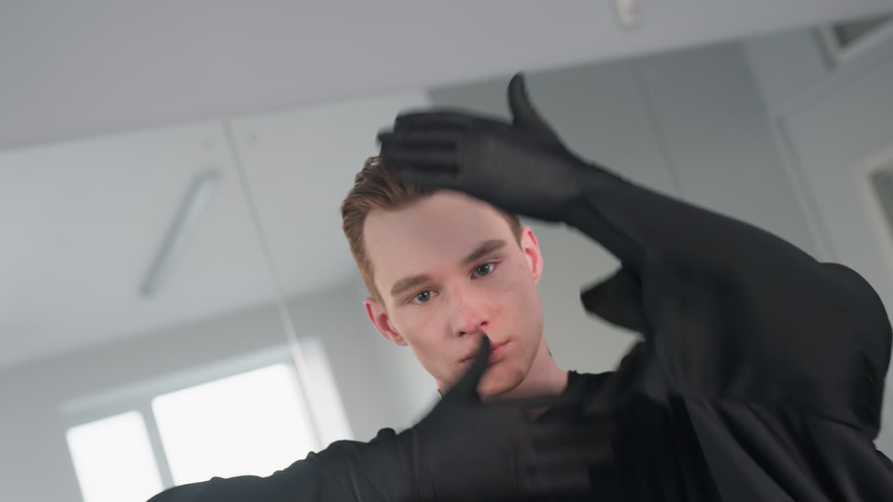 young man in dark cloak making expressive hand gesture around face with gentle smile, performing stylish dance poses in front of large mirror in studio environment with reflective backdrop