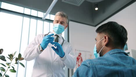 Close-Up-Portrait-Of-Handsome-Experienced-Senior-Male-Doctor-Virologist-In-Medical-Mask-Injecting-Covid-Vaccine-To-Man-Rear-Of-Young-Hindu-Patient-Sitting-In-Medical-Center-Receive-Vaccine-Vaccination