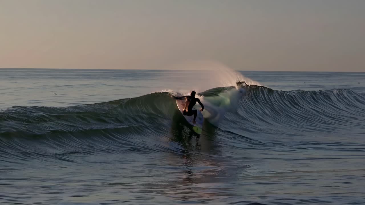 A surfer balances on a board in the ocean at sunset. Captured from a low angle, the video conveys