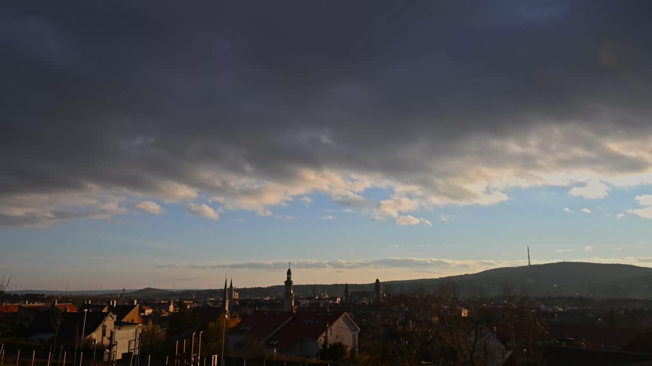Time lapse video of a warm sunset illuminates a small city with church towers and rooftops, while dramatic clouds moving across the sky. The distant hills and soft golden light create a calm scene