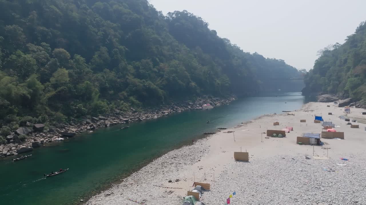 Dawki, Meghalaya, India — barcos a la deriva sobre aguas cristalinas de río turquesa flanqueadas por laderas boscosas. La ribera arenosa con estructuras dispersas bordea la orilla opuesta bajo un cielo nublado