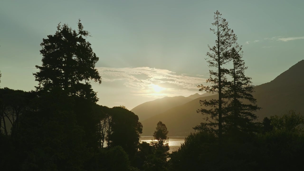 Plano médio cinematográfico através das árvores em direção a um lago de montanha ao pôr do sol. A luz dourada do sol desponta acima do cume enquanto um pássaro voa pelo enquadramento, criando uma cena de verão tranquila.
