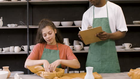 young woman wrapping handicraft ceramics with paper while her male colleague writing on clipboard in the pottery shop 1