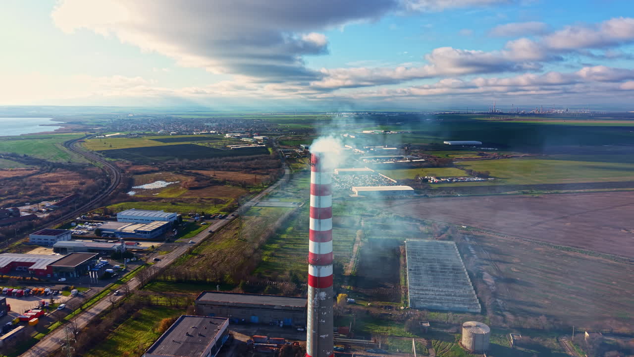 Il fumo sale da un'alta ciminiera in un sito industriale vicino a un campo e a uno specchio d'acqua sotto un cielo nuvoloso durante il giorno