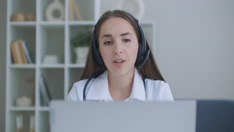 female medical assistant wears white coat headset video calling distant patient on laptop. doctor talking to client using virtual chat computer app. telemedicine remote healthcare services concept.