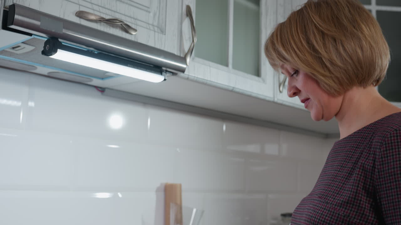 Side profile of housewife thoughtfully preparing dinner in brightly lit cozy kitchen, featuring white glossy tiles, cooking utensils, modern cabinetry, neatly arranged countertop items