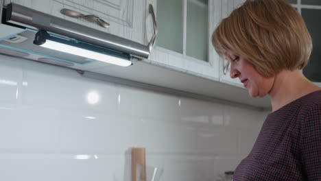 side profile of housewife thoughtfully preparing dinner in brightly lit cozy kitchen, featuring white glossy tiles, cooking utensils, modern cabinetry, neatly arranged countertop items