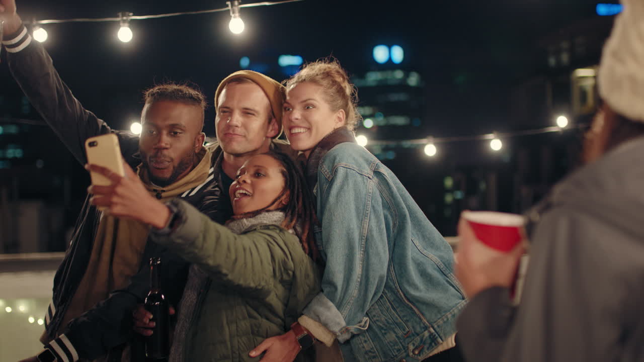 행복한 여성이 스마트 폰을 사용하여 친구들과 그룹 사진을 찍고 재미있는 얼굴을 만들고 소셜 미디어에서 주말 옥상 파티를 공유하고 밤에 재미를 느끼고 있습니다.