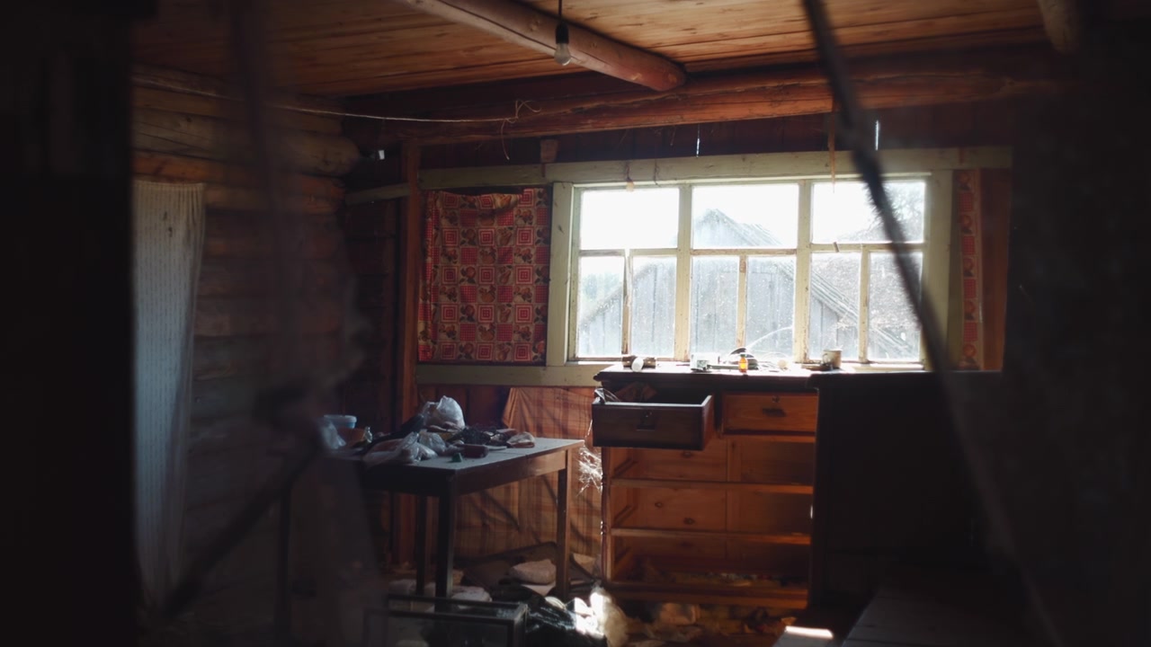 Interno di baita rustica illuminato dal sole con finestra, vecchia cassa di legno accanto a una tenda a fantasia, attrezzi sparsi.