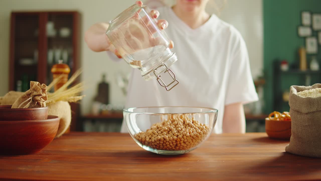 Person pouring chickpeas into a glass bowl on a wooden table