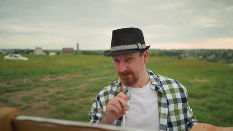 a close-up of a painter wearing a hat and a checkered shirt, standing in a grass field as he playfully scratches his nose with a paintbrush. the background shows a rural landscape under a cloudy sky