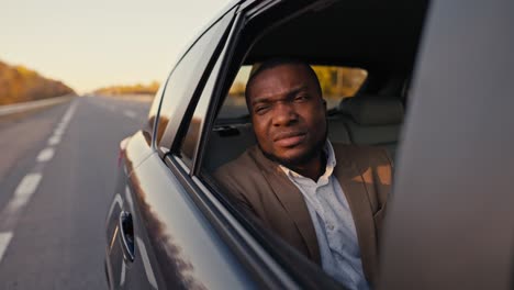 A-suspicious-Black-man-with-a-dissatisfied-face-in-a-brown-suit-opens-the-rear-window-of-the-car-and-looks-at-the-roadway-during-his-trip-outside-the-city.-Dissatisfied-pensive-male-businessman-looking-out-the-window