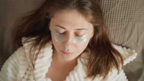 woman lies on soft sofa surface listening to pleasant music in room. lady puts relaxing patches under closed eyes enjoying pleasant leisure time on day