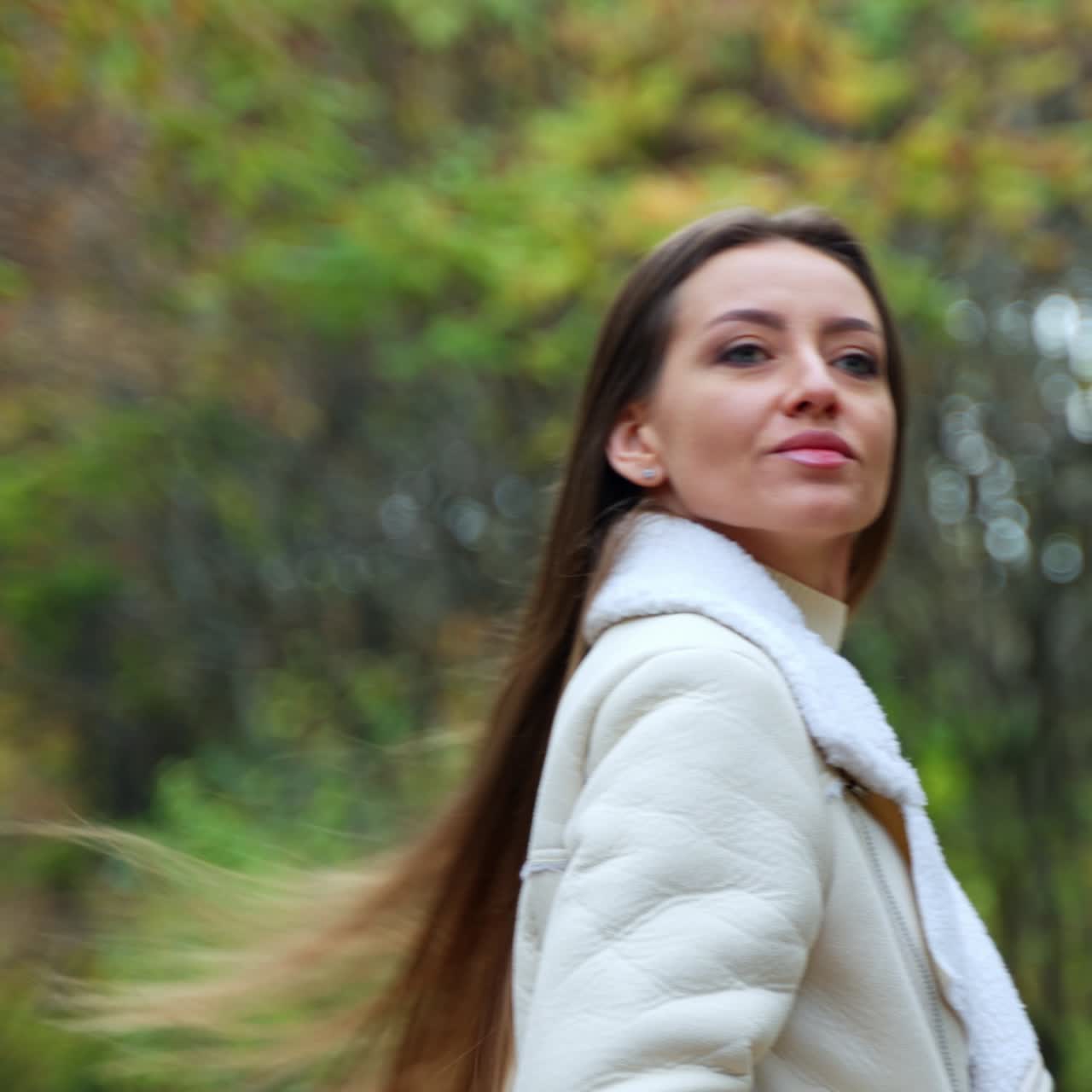 Smiling brunette in light warm jacket walking in the nature. Girl taking a stroll in the park in autumn. Blurred backdrop