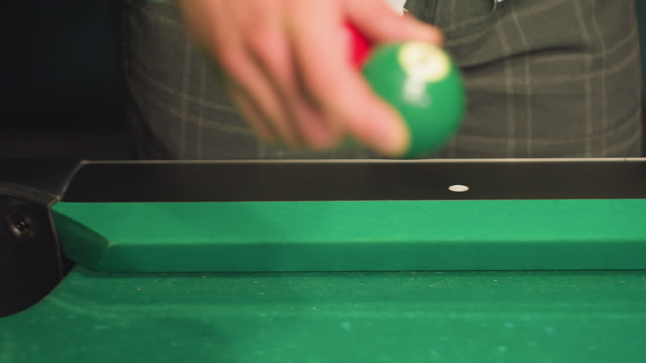 Person in white shirt and grey trousers retrieving green ball from green pool table hole. Close-up shot captures hand movement, texture of pool table, and game interaction in dimly lit
