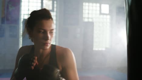 Slow-Motion-shot:-beautiful-female-boxer-in-gloves-hitting-boxing-bag-with-her-fist-while-training-in-a-dark-fitness-studio-with