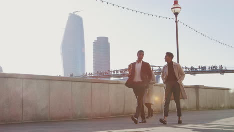 Two-business-colleagues-walking-by-the-Thames-riverside-near-Millennium-Bridge-in-the-city-of-London-talking,-man-holding-a-takeaway-coffee,-lens-flare