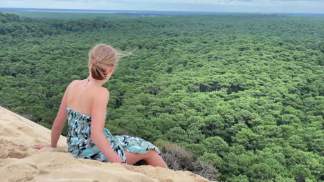 A-young-girl-is-watching-a-large-forest-on-the-horizon,-sitting-on-a-dune-in-south-west-France,-dunes-of-pilat