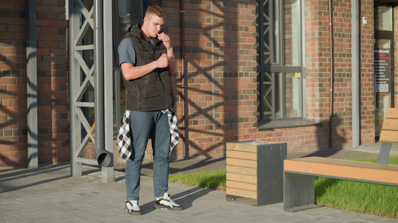 young boy standing outdoors beside wooden bench brings cigarette to mouth then decides against lighting it and tosses it into nearby waste bin under sunlight near brick wall and metal frame structure