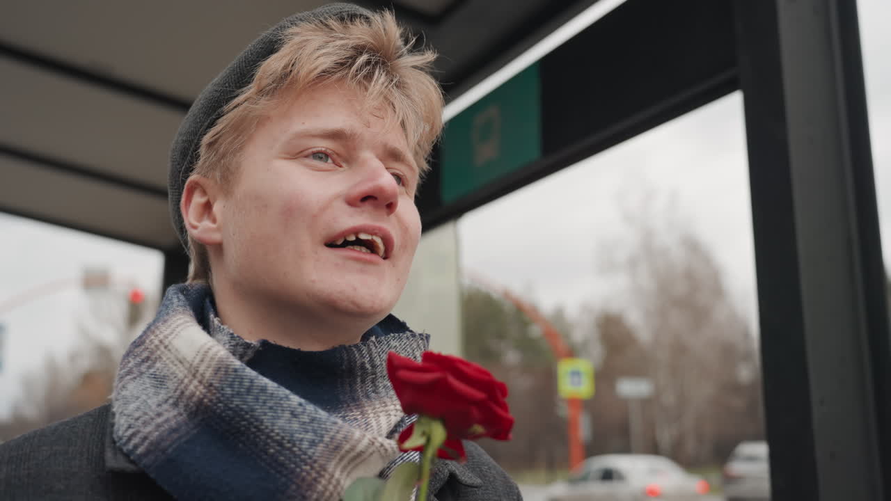 Nuori valkoihoinen mies tarjoaa ruusua, luoja harjoittelee vuorosanoja bussipysäkillä, ilmeikäs hymy ja eleet, huivi ja pipo, aito urbaani hetki, leikkisä koe-esiintymisen tunnelma ja hiljainen optimismi.