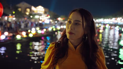 Woman-in-yellow-Ao-Dai-by-Hoi-An-river-at-night,-vibrant-lantern-lights,-reflecting-on-water,-contemplative-mood