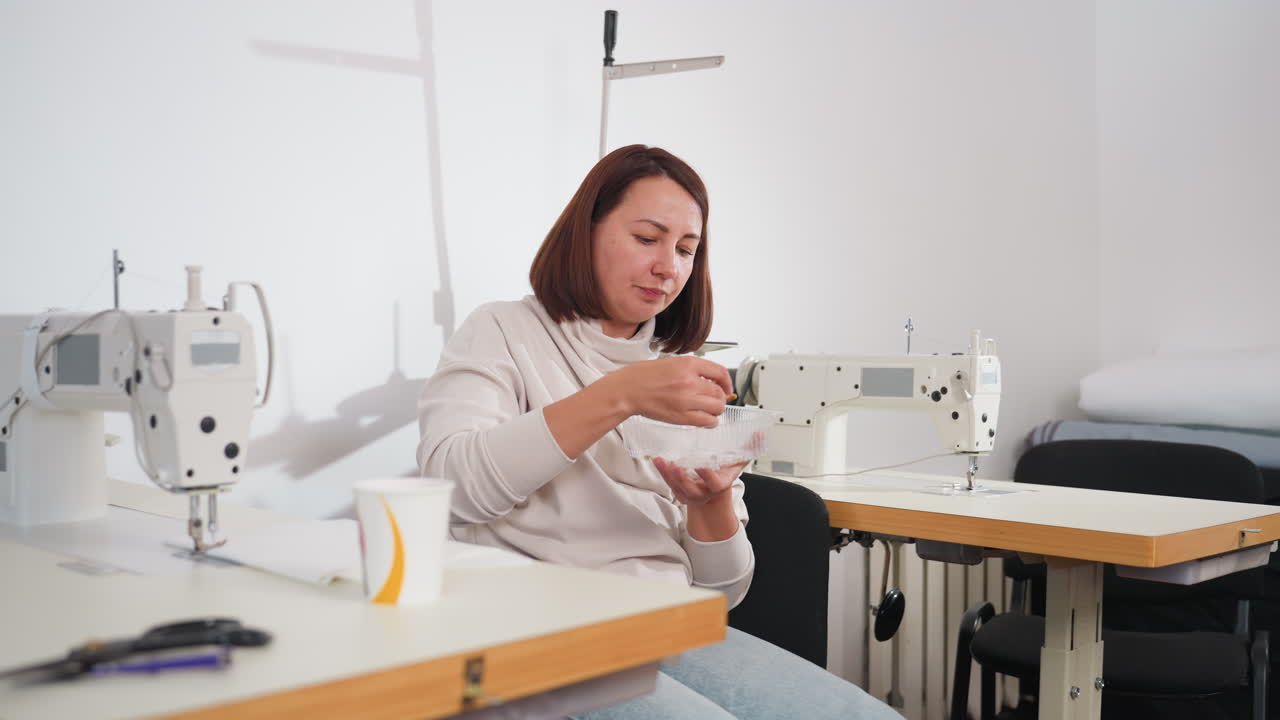 Female seamstress wiping fingers with tissue after eating pizza while seated between sewing machines, cup of tea and scissors on table, relaxed indoor workspace with white background atmosphere