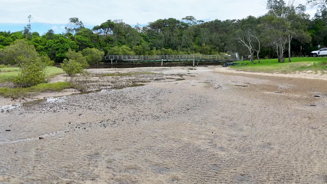 Tidal water rapidly fills sandy estuary under overcast daylight, camera moves forward toward footbridge
