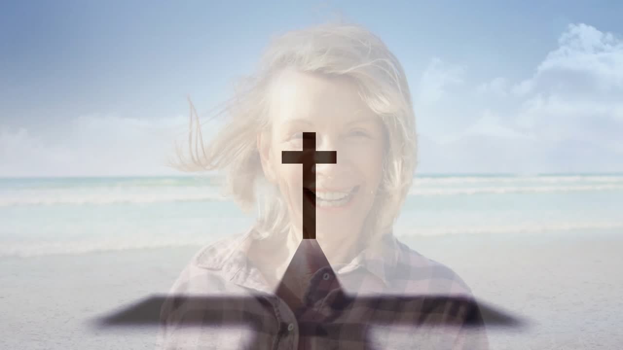 Senior woman turning and smiling at shore, gusts causing cross and book overlay growing for faith