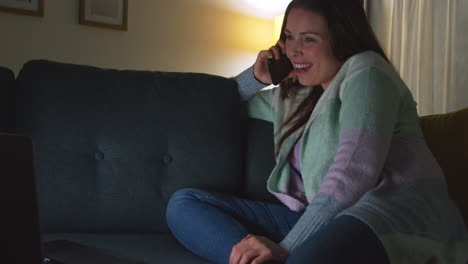 Smiling-Woman-Sitting-On-Sofa-At-Home-At-Night-Talking-On-Mobile-Phone-And-Watching-Movie-Or-Show-On-Laptop-8