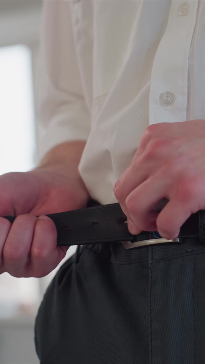 Primer plano de un hombre con atuendo formal ajustándose el cinturón. El hombre viste camisa blanca con mangas remangadas y pantalones negros. El escenario es un espacio interior luminoso con grandes ventanas o paredes de color claro.