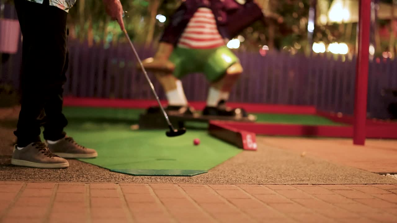 A man play mini golf under warm lighting, focusing on a putt attempt. The scene captures playful energy and concentration
