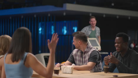 A-multi-ethnic-group-of-friends-sits-at-a-table-together-in-a-bowling-alley-and-talks-give-five-to-a-joyful-man-jumping-for-joy-and-celebrating-victory-and-a-knocked-out-clash.