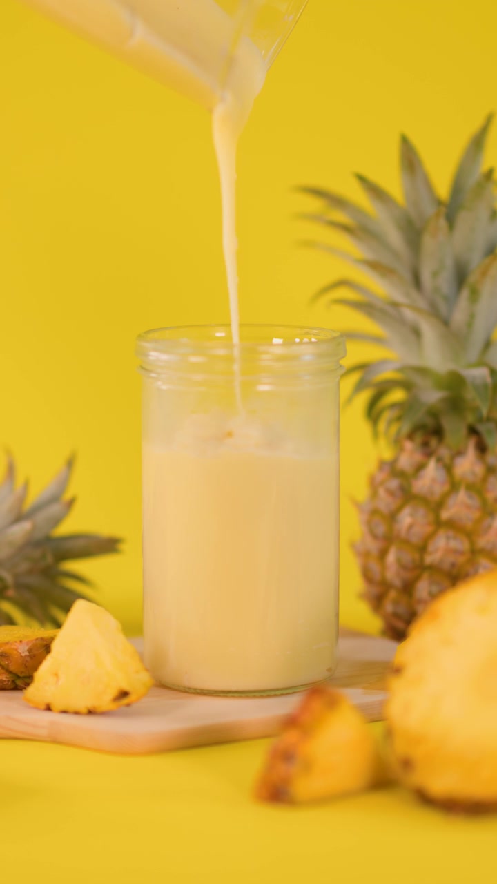 Frullato di ananas versato in un barattolo di vetro, circondato da frutta fresca, sfondo giallo brillante, illuminazione soffusa.