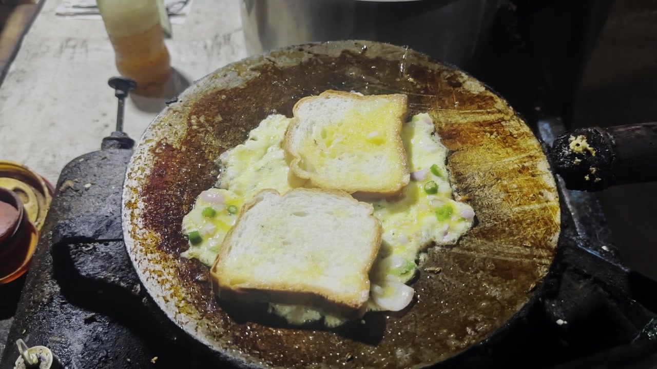 Omelet roti gaya jalanan disiapkan dengan telur, roti, bawang, dan cabai hijau di atas tawa panas oleh penjual lokal
