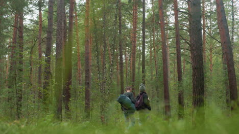 travelers pause in dense forest after dropping bags as man places hand on waist and scans surroundings thoughtfully, surrounded by tall trees and natural greenery in calm woodland setting