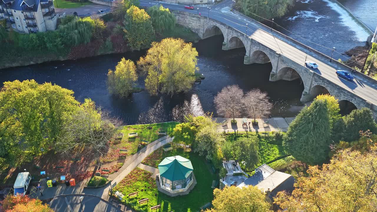 Aerial view of Riverside Bandstand, green park with pathways, mature trees and historic stone bridge over river in Wetherby. Autumn landscape with golden foliage and tranquil water