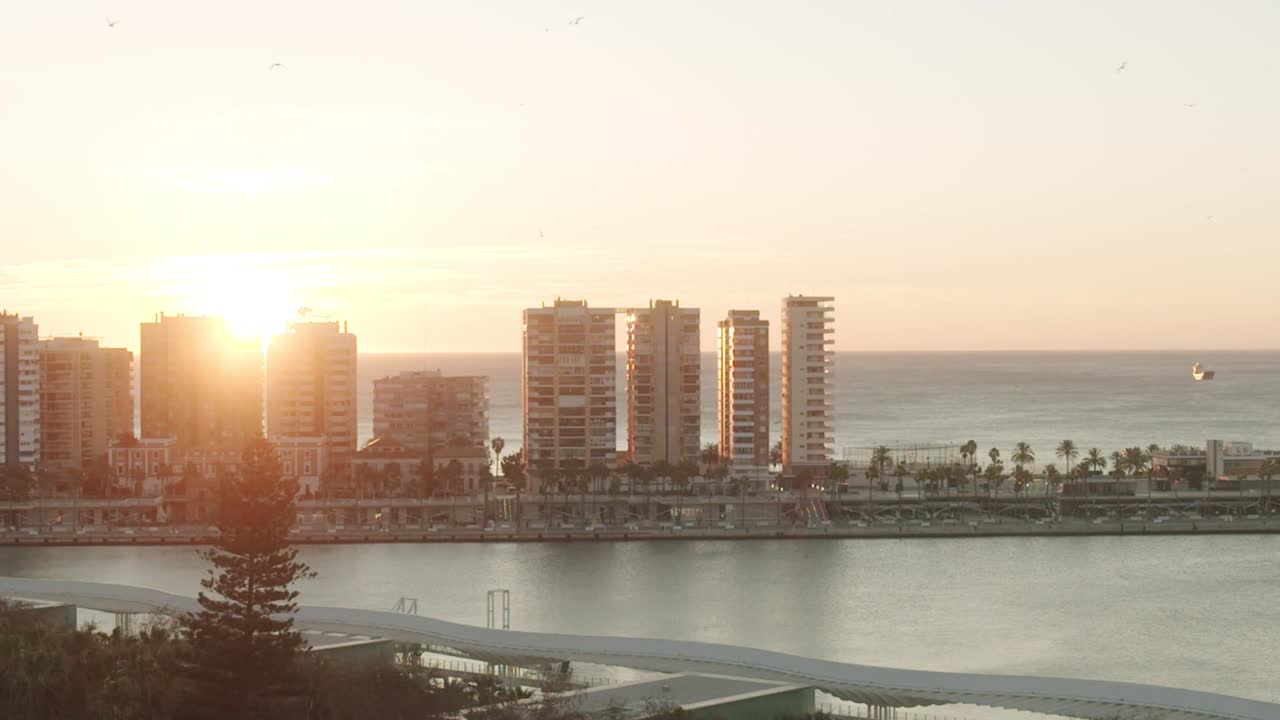 Glowing orange sun sets behind high rising apartments of harbor coastline of Malaga City, Spain