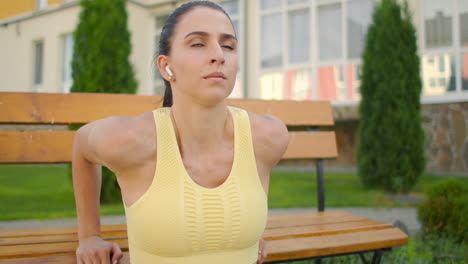 una mujer joven con auriculares realiza flexiones en un banco en un parque de la ciudad en cámara lenta. entrenando a una mujer joven en un banco con el telón de fondo de la ciudad y las casas